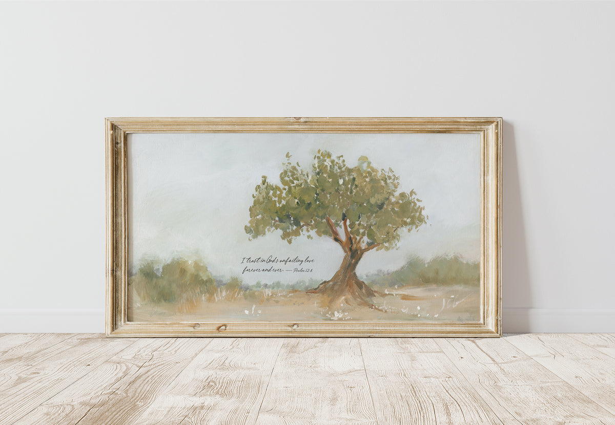 A framed watercolor landscape print of a solitary tree in a field, leaning against a white wall on a light wood floor. The artwork features a light beige wood frame and includes a small cursive quote  Psalm 52:8 “I trust in God’s unfailing love forever and ever.”